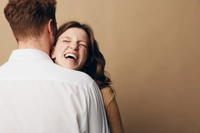 Happy woman with brown hair laughs joyfully while hugging a man in white shirt. Warm beige background creates cozy atmosphere. Concept of love, happiness, and intimate relationship.