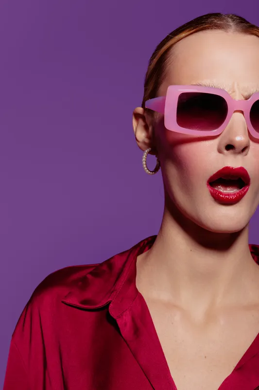 Young woman in pink sunglasses and red shirt shows surprised expression on purple background. Fashion portrait with bold makeup and modern accessories. Concept of style and attitude.