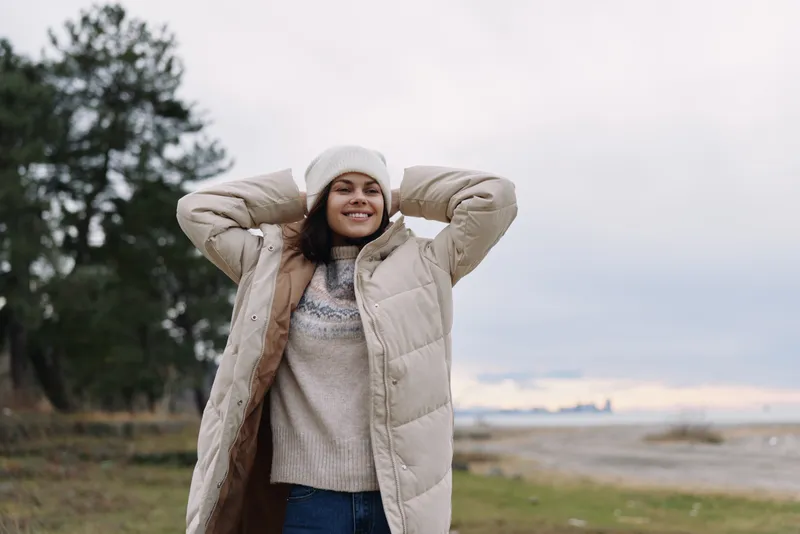 Woman wearing a warm beige winter coat and beanie standing outdoors with hands behind her head, smiling, in a chilly landscape by a lake and trees under a cloudy sky.