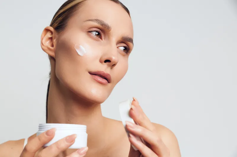 A young woman applies white face cream on her cheek while holding a jar of moisturizer in a bright studio setting. Concept for skincare, beauty routine, and healthy skin care.