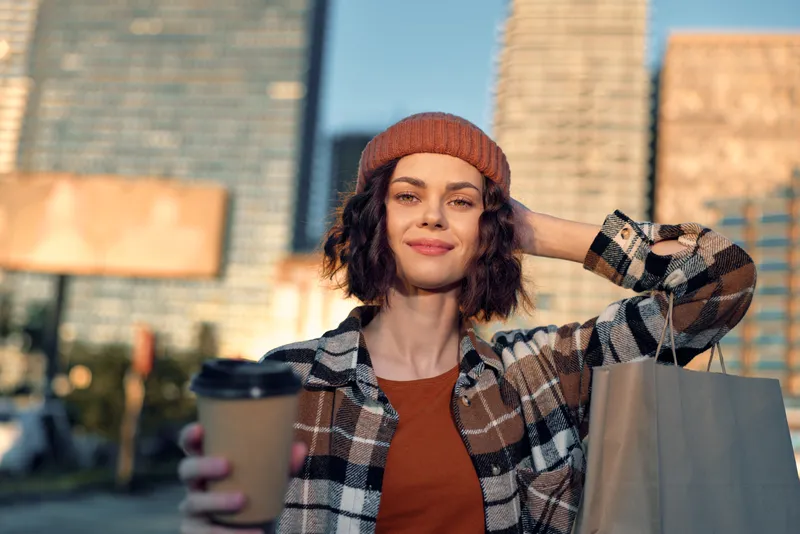 Woman holding coffee and shopping bag in city at golden hour, beanie and plaid jacket, smile and urban lifestyle portrait with authenticity, candid lifestyle, mindful living, emotional storytelling.