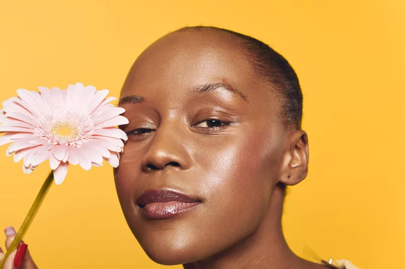 Close-up of a confident black woman with dark skin holding a pink flower near her face on a yellow background. Concept of beauty, skincare, natural glow, and wellness.