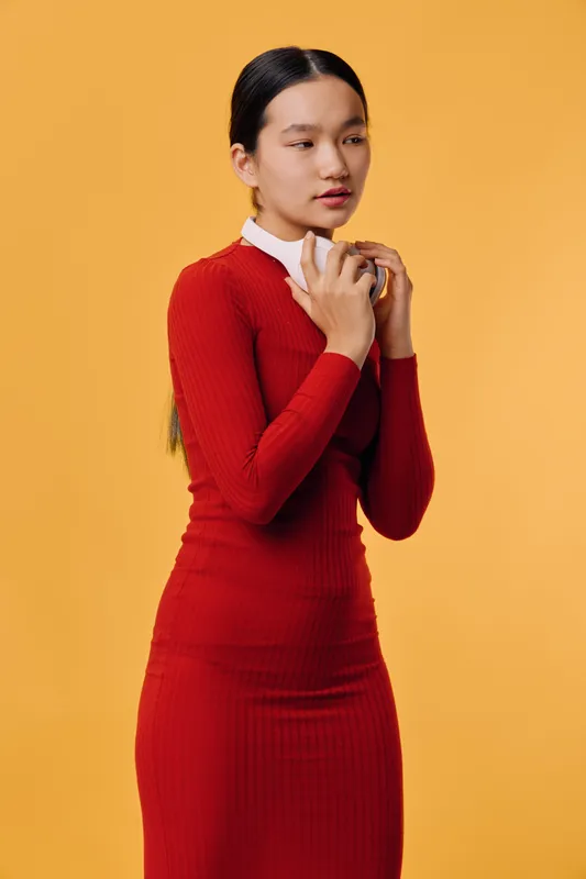 Young woman in red dress standing with clasped hands against orange background, thoughtful expression and long dark hair tied back in studio portrait. Fashion, elegance and contemplation.