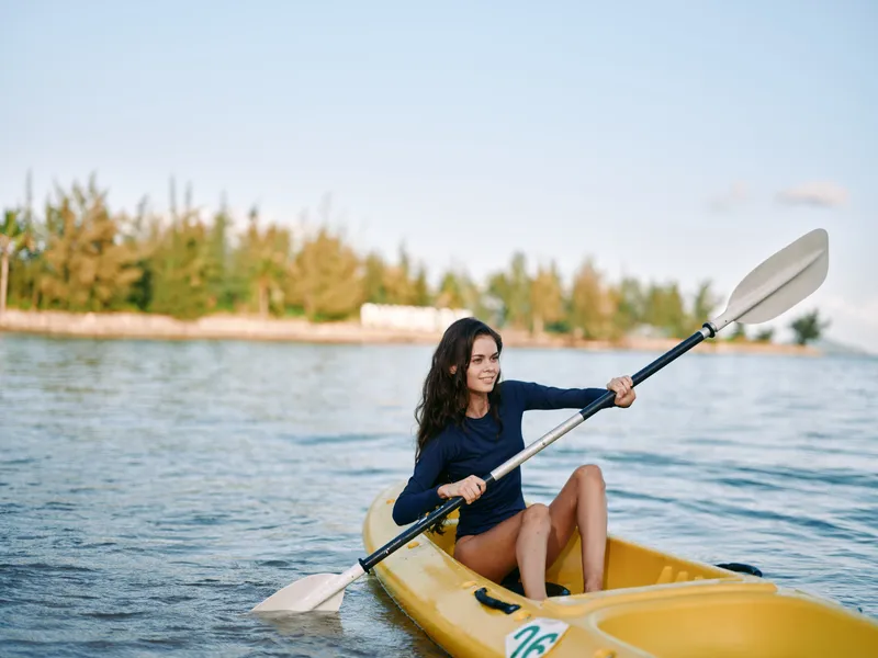 Young woman paddles a yellow kayak on calm water, sunlight glinting off the surface as she smiles and calmly navigates with a double paddle near a tree-lined shore.