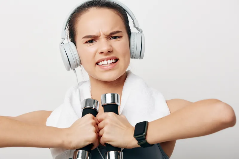 Female fitness enthusiast lifting dumbbells with headphones and a towel draped over her shoulders, intense expression, indoor gym setting, focus on strength and determination during a workout.