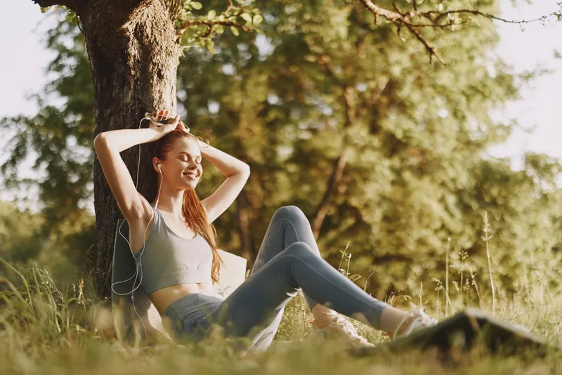 Young woman enjoying music under a tree in a green park, wearing casual clothing, exuding joy and relaxation, perfect for themes of wellness and leisure