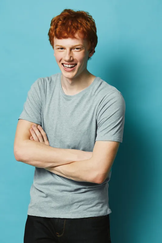 Young man with red hair and freckles wearing gray t shirt smiling confidently with arms crossed standing against blue background. Casual style and relaxed posture with happy expression.