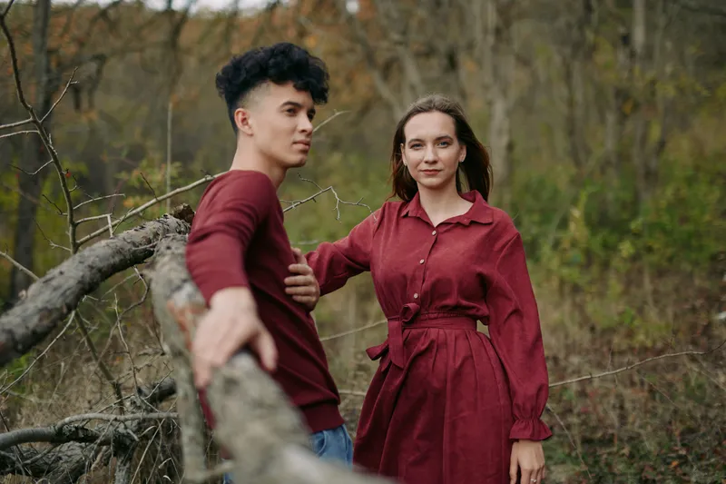 Young couple in coordinated burgundy outfits pose naturally by a fallen tree in a quiet woodland, their relaxed expressions conveying an authentic connection and quiet credibility in a candid outdoor