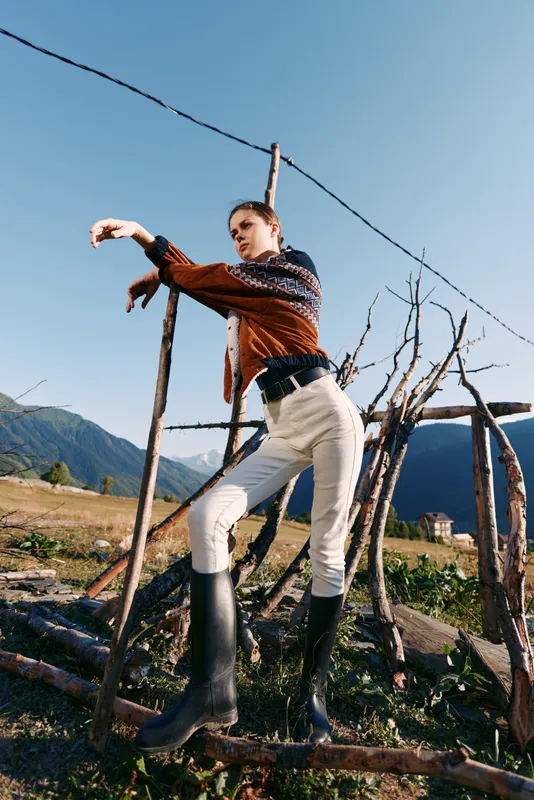 Woman model in chunky sweater and tall boots poses by rustic fence in open countryside with distant mountains and blue sky, fashion portrait capturing outdoor lifestyle and natural texture