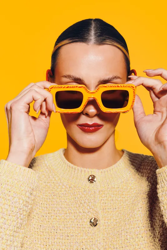 Young woman with slicked hair wearing yellow sunglasses and a beige sweater poses against bright yellow background. Concept of fashion, style, summer, and confidence.
