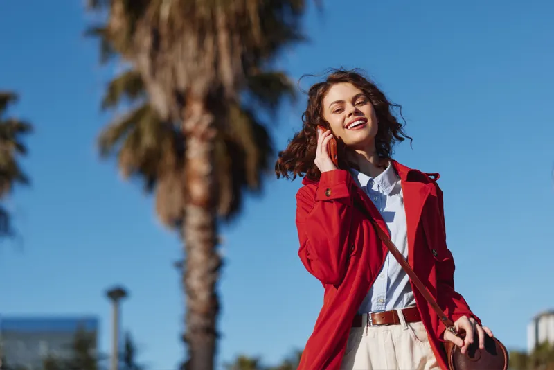 Young woman with curly hair wearing a red coat smiles happily while walking outdoors on a sunny day with a clear blue sky and palm trees in the background.
