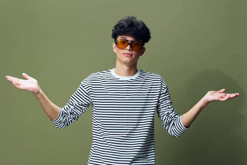 young man with curly hair wearing sunglasses and striped shirt showing gesture of uncertainty against olive background, casual style, confident expression