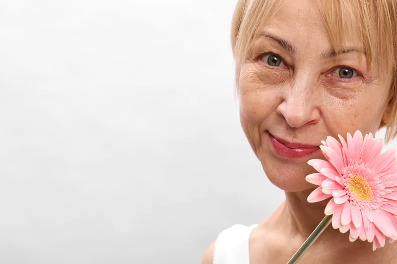 Smiling woman portrait in closeup, holding a pink gerbera daisy near the face, soft natural light, white background, calm skincare beauty mood, gentle expression