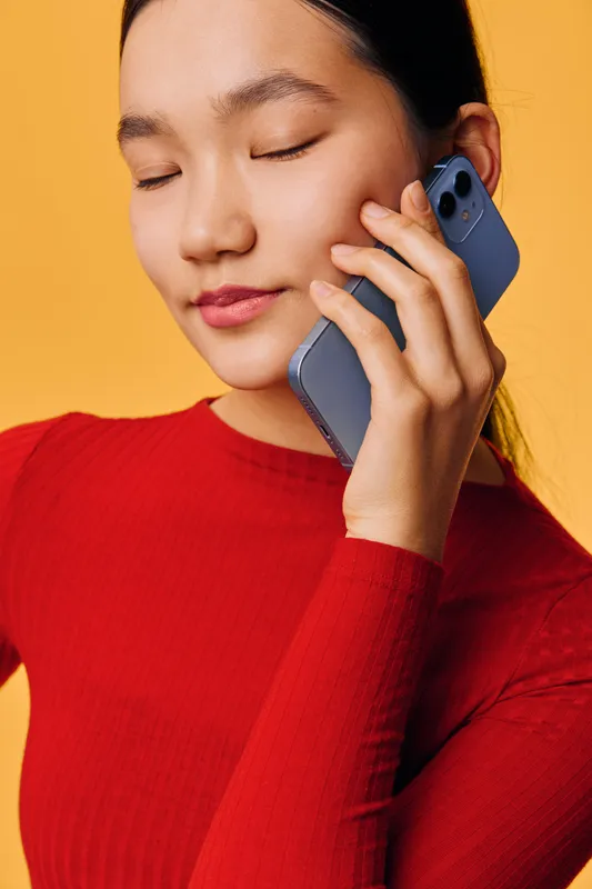 Young woman with closed eyes holding smartphone near ear for call or conversation with calm expression. Asian female in red top against yellow background for communication concept.
