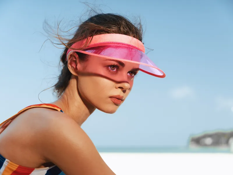 Portrait of a young woman wearing a pink translucent visor at a sunny beach. Her face is focused, hair swept by sea breeze, warm light highlights smooth skin and a relaxed, athletic vibe.