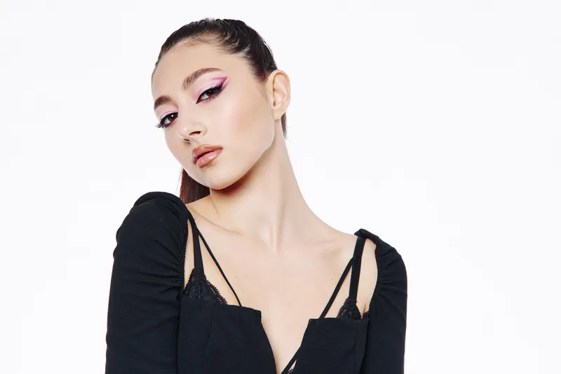 Young woman with flawless skin wearing bold pink eyeliner posing confidently in a minimalist black outfit in front of a white background. Beauty fashion concept.