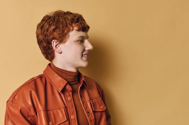 Young man with red hair and fair skin wearing a brown leather jacket and turtleneck standing against beige background in profile view with slight smile.