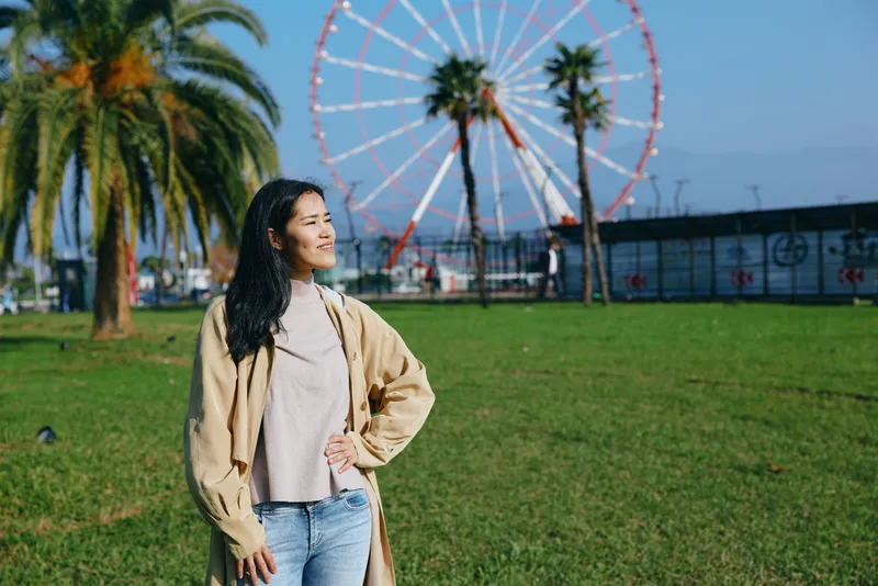 Woman standing in a sunny park with a large ferris wheel in the background, wearing a cozy coat and sweater, enjoying a calm outdoor moment, fashionable, relaxed, and carefree.