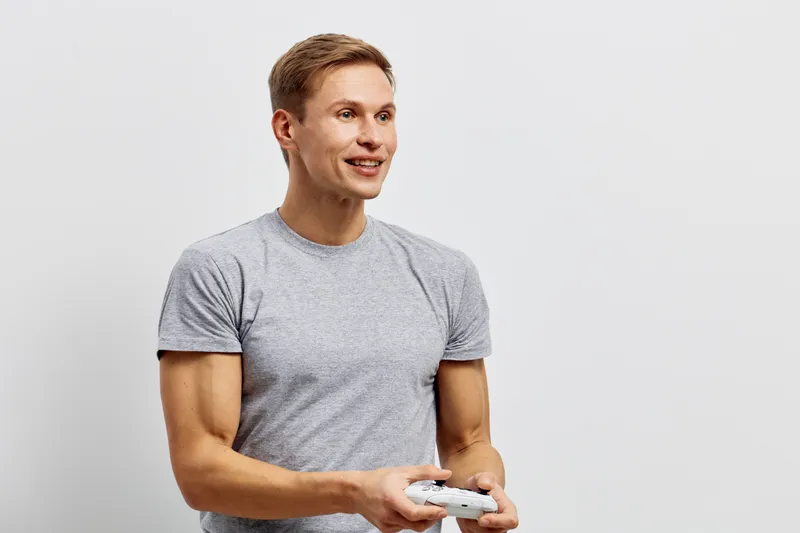 Smiling young man with short hair wearing casual gray t-shirt holding game controller look friendly positive isolated on plain white background studio portrait. People lifestyle concept