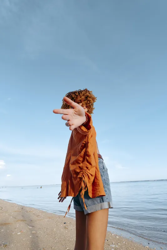 Young woman with curly hair wearing orange sweater and denim shorts standing on sandy beach by calm sea under clear blue sky, casual summer outfit, carefree mood