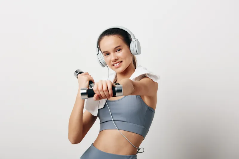 Young woman fitness portrait in studio, sport concept with headphones, smiling while holding dumbbells and towel on shoulder against a clean white background, active confident pose.
