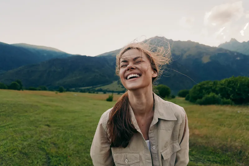 A happy woman with long hair smiles outdoors in a green field with mountains in the background. Natural light highlights her joyful expression and casual outfit.