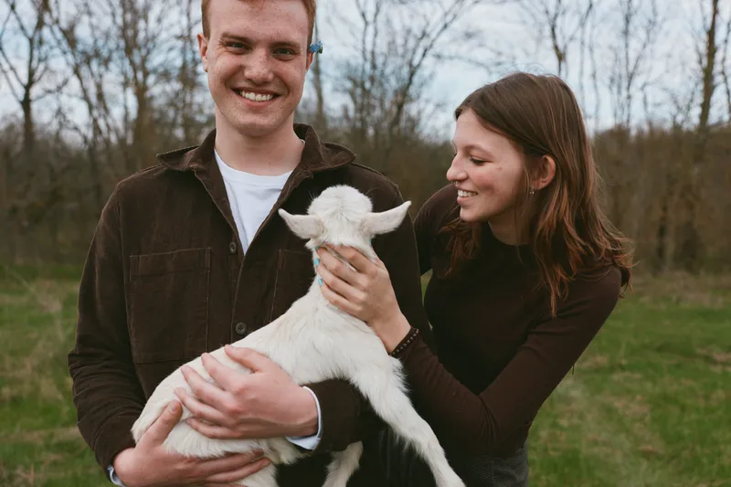 A happy couple outdoors in a field gently holds a young goat kid, smiles, and shares a calm moment, showcasing rural lifestyle and tender animal interaction, connection, and warmth against a natural