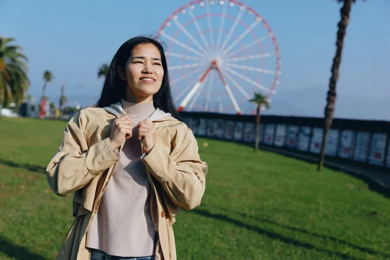 Smiling woman in a casual jacket stands outdoors near a boardwalk with a large ferris wheel in the background, sunny day, relaxed mood, travel and leisure scene for travel