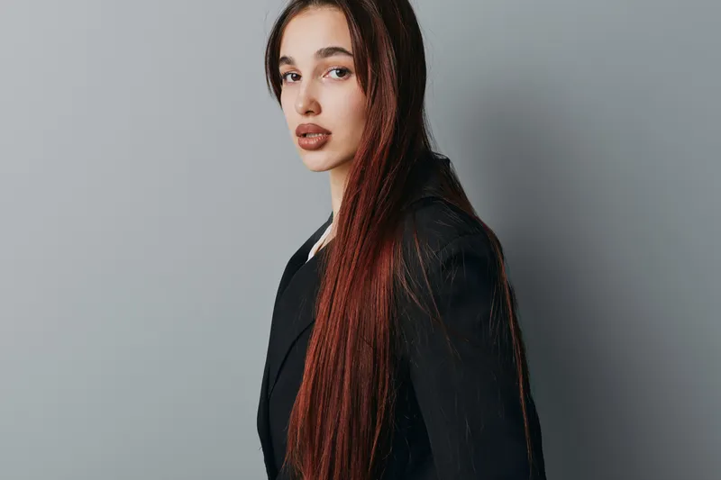 Young woman with long straight hair wearing black blazer posing and looking at camera with neutral expression on gray background in studio portrait. Female model with natural makeup.