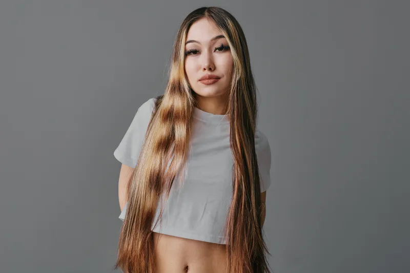Young woman with long straight hair and casual white crop top posing on neutral gray background for beauty and fashion portrait. Female model looking at camera with calm expression.