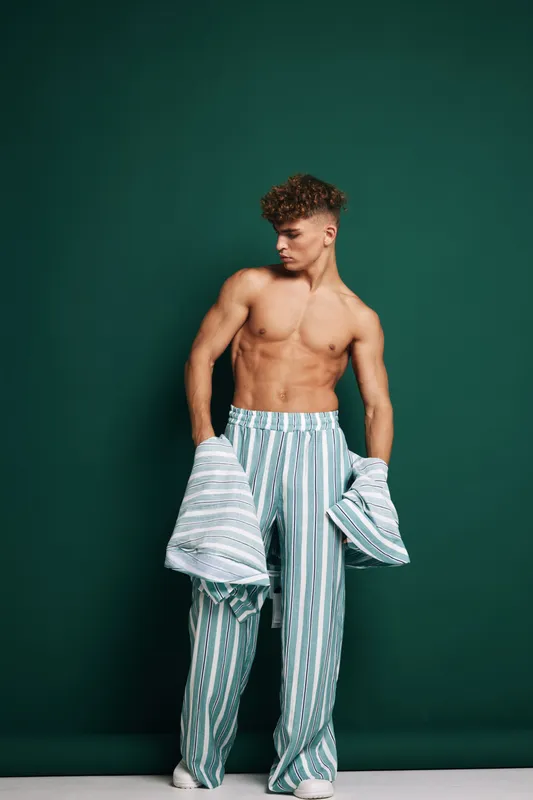 Man standing with relaxed pose looking sideways holding matching striped jacket and pants. Muscle body, haircut hairstyle, casual fashion, isolated green background, front view, shirtless young adult.