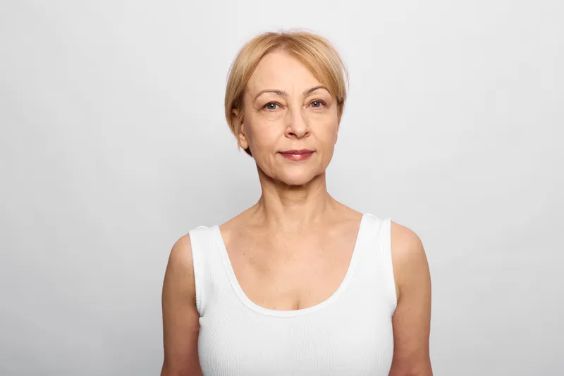 Woman portrait, mature beauty and wellness concept in studio, gray background. Older blonde woman in white tank top, calm expression, soft light closeup