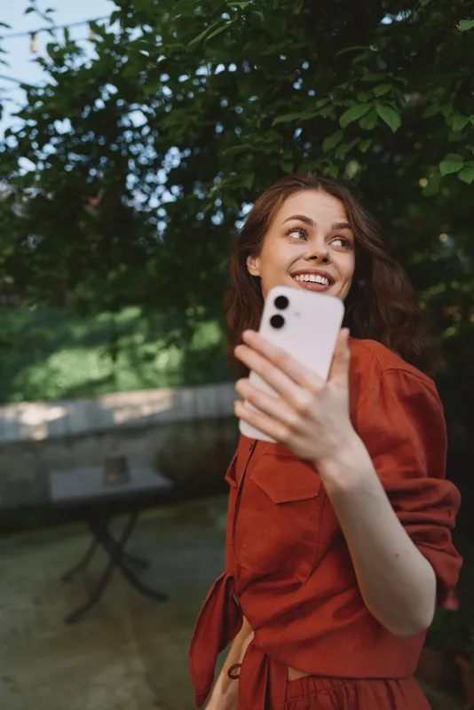 Smiling woman with wavy hair taking a selfie outdoors, wearing a rust red dress, lush greenery in the background. A candid lifestyle moment focused on joy, self expression, and photography
