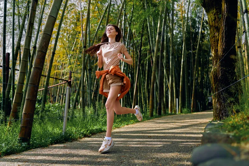 Woman running along a bamboo forest path, fitness jog and outdoor exercise with a smile, casual shorts and sneakers, sunlight, motion and healthy lifestyle on a scenic trail.