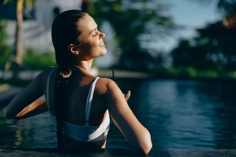 A young brunette woman in a white swimsuit relaxes at the pool edge, smiling with her back to the camera as sunlit water glitters in the background.