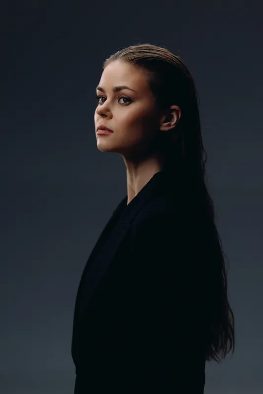 Profile of a young woman with long hair wearing a black outfit against dark background, showing serious expression and elegant style.