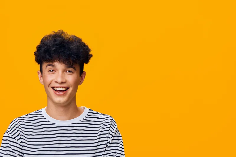smiling man with curly hair wearing striped shirt against bright orange background, happiness, youth, casual style, positive emotion, studio lighting