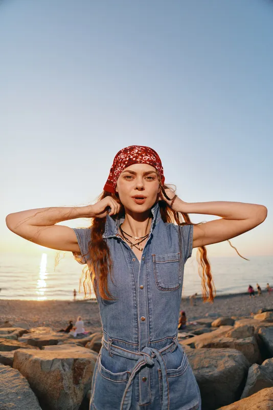 lifestyle woman in modern boho-western denim street style adjusting red headscarf at sunset beach with film color tones and relaxed mood