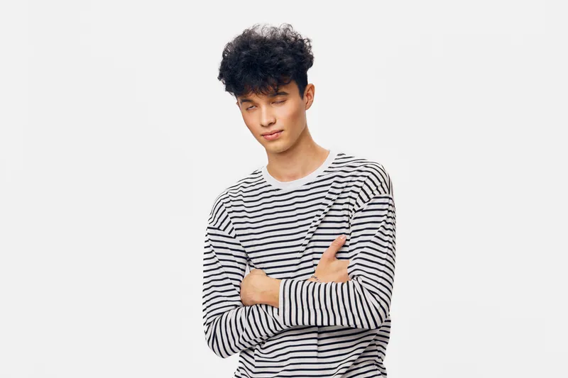 young man with curly hair wearing striped shirt standing with crossed arms, closed eyes and relaxed expression on white background, casual style portrait