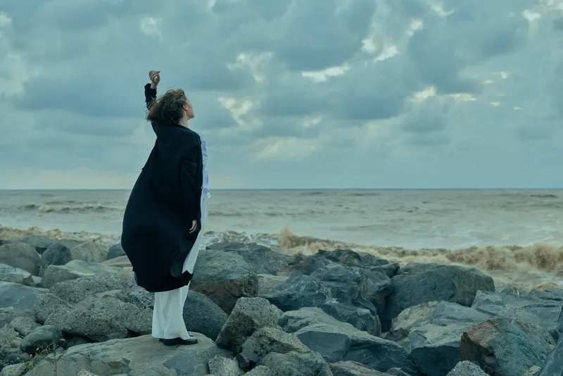 Woman on a rocky shore raises a clenched fist toward the turbulent sea under a heavy cloudy sky, capturing resilience, determination and a dramatic coastal mood that conveys courage against the