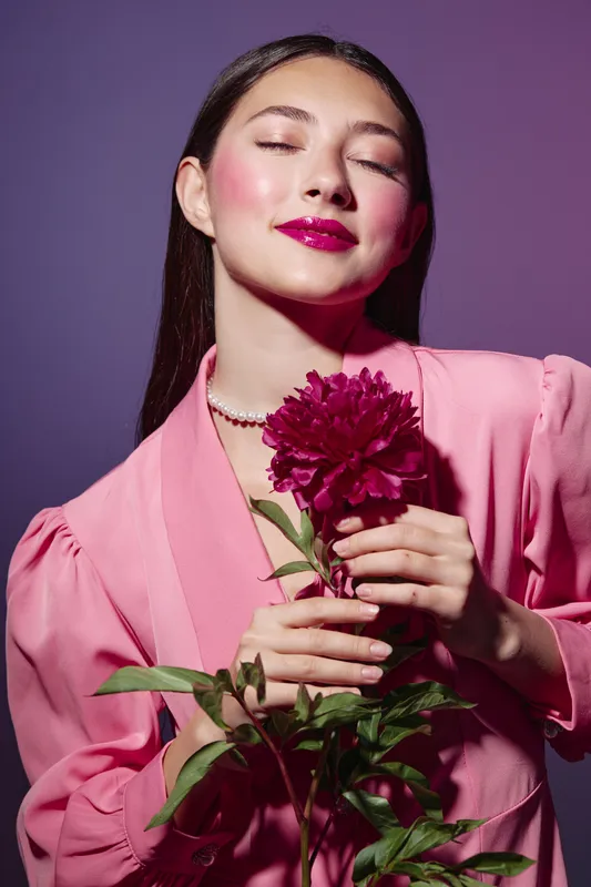 Young woman with long dark hair wears pink stylish attire holding a bold magenta flower with closed eyes. Studio portrait on purple background concept for beauty fashion creativity.