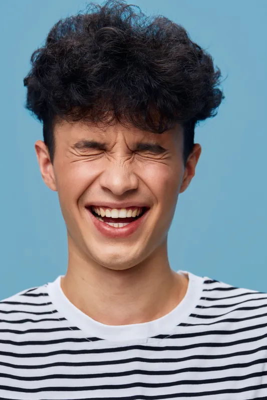 smiling man with curly hair wearing striped shirt laughs joyfully with closed eyes against light blue background, portraying happiness and lively energy in youthful style.