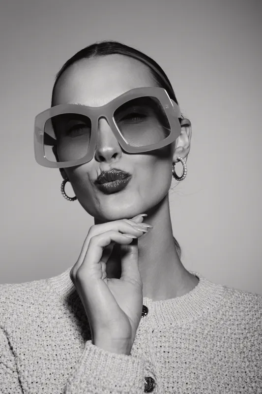 Stylish white woman with pale skin wearing large sunglasses and hoop earrings poses with hand near face in black and white studio portrait. Concept of fashion and elegance.