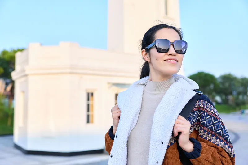 Sunglasses, woman, coat, scarf outdoors under bright daylight beside white architecture, fashion forward, confident pose, casual urban style photography for social