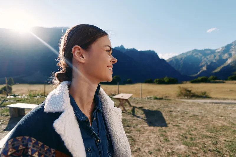 Woman in jacket smiles in sunlight against mountains and open countryside, profile portrait capturing a happy young traveler enjoying nature, scenic field and relaxed outdoor mood.
