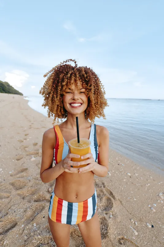 Woman, beach, ocean, summer, smiling, holding a smoothie with a straw, curly hair, striped swimsuit, sunlit sand and ocean waves behind, outdoor vacation, refreshment concept