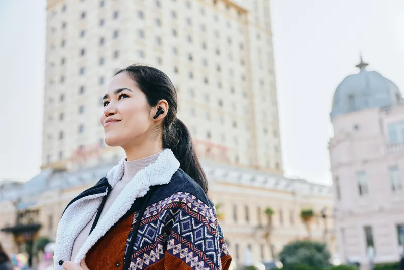 Woman in stylish coat and scarf stands in a sunlit cityscape, headphones on, smiling, embodying urban fashion, travel vibe, street style, and confident pose amid tall architecture and bright daylight
