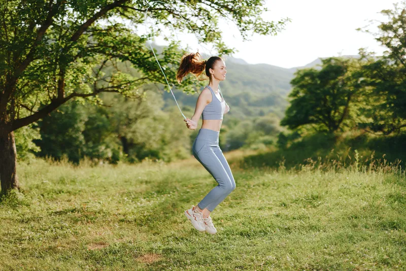 Active young woman skipping rope outdoors in lush green nature, embodying vitality and fitness in stylish sportswear