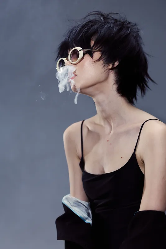 Stylish woman in sunglasses exhaling smoke with casual black outfit and edgy hairstyle on gray background