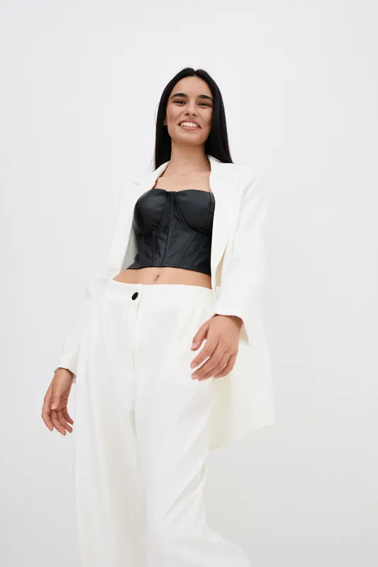 Smiling woman in white suit and black top stands confidently against white background, showcasing modern fashion with a relaxed and positive attitude.